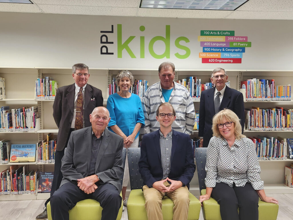 Photo of seven individuals, three sitting, four standing in front of bookshelves